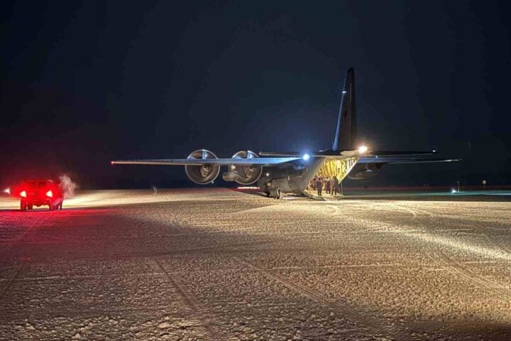 An RNZAF C-130J Hercules sits on a groomed ice runway at McMurdo Station under dark Antarctic skies as crew members stand by the open rear ramp.