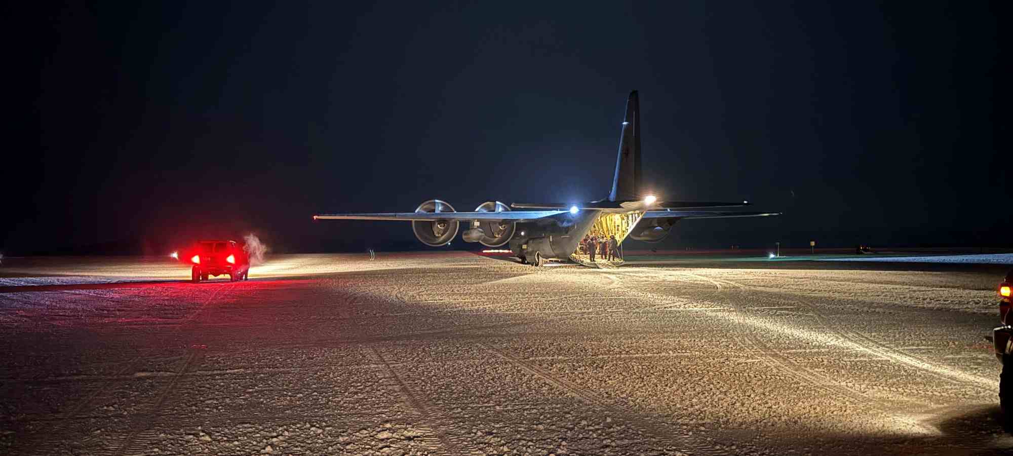 An RNZAF C-130J Hercules sits on a groomed ice runway at McMurdo Station under dark Antarctic skies as crew members stand by the open rear ramp.