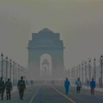 Pedestrians and uniformed guards walking along a foggy Rajpath with India Gate looming through thick haze at dawn in New Delhi.
