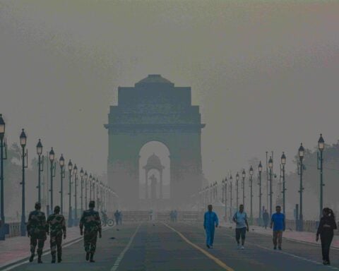 Pedestrians and uniformed guards walking along a foggy Rajpath with India Gate looming through thick haze at dawn in New Delhi.