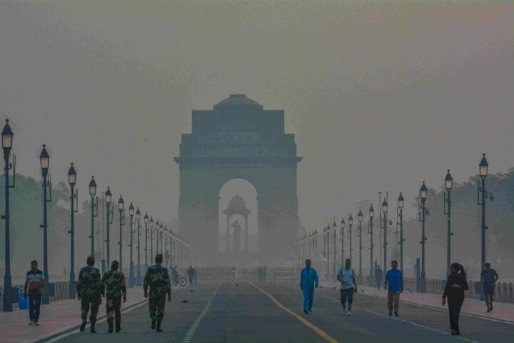 Pedestrians and uniformed guards walking along a foggy Rajpath with India Gate looming through thick haze at dawn in New Delhi.