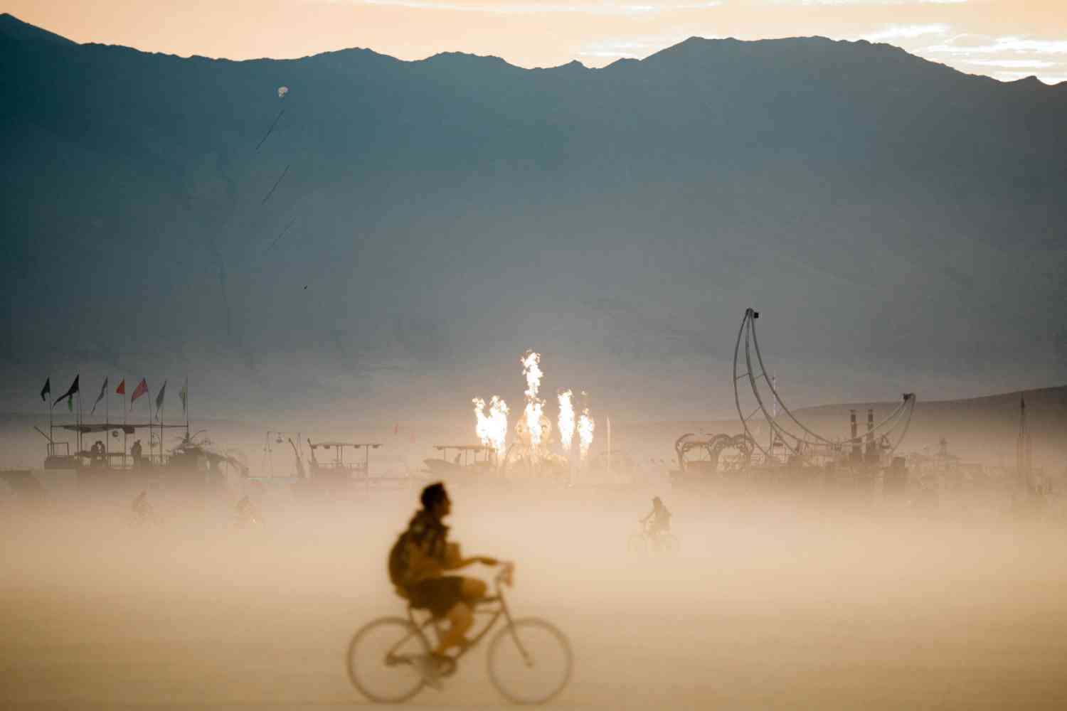 Person riding a bicycle across the dusty playa at Burning Man Festival, with fire art installations and flags in the desert background.