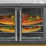 Oster French Door Countertop Oven model TSSTTVFDMAF with stainless steel finish, featuring center-opening glass doors and control knobs on the right side, shown cooking multiple pizzas on two racks.