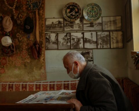 Elderly man wearing a face mask reading a newspaper at a table in his home, with vintage photographs and decorative plates displayed on the wall behind him.