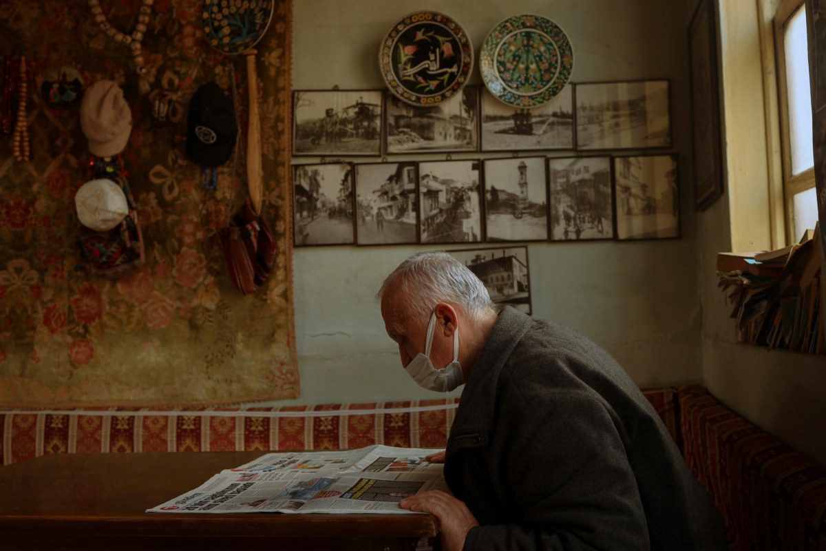 Elderly man wearing a face mask reading a newspaper at a table in his home, with vintage photographs and decorative plates displayed on the wall behind him.