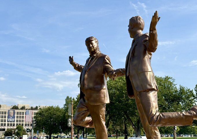 Bronze statues of two suited figures holding hands and lifting one leg, installed on marble pedestals with the U.S. Capitol building in the background.