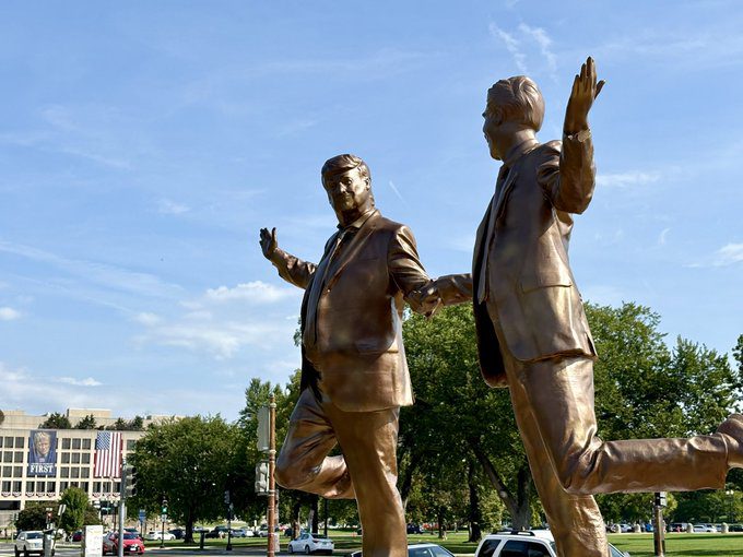 Bronze statues of two suited figures holding hands and lifting one leg, installed on marble pedestals with the U.S. Capitol building in the background.