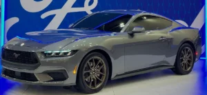 Side-front view of a silver 2024 Ford Mustang coupe with dark wheels displayed at an auto show, featuring the redesigned front end with signature tri-bar LED headlights characteristic of the seventh-generation model.