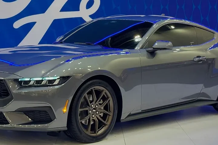 Side-front view of a silver 2024 Ford Mustang coupe with dark wheels displayed at an auto show, featuring the redesigned front end with signature tri-bar LED headlights characteristic of the seventh-generation model.