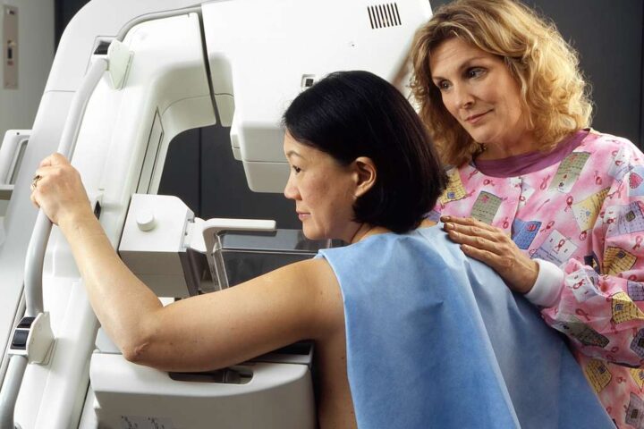 A healthcare professional assists an Asian woman positioning herself for a mammogram screening, demonstrating proper breast cancer screening procedure.