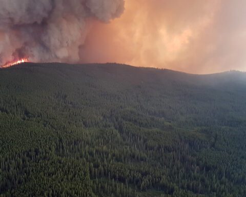 Aerial view of the Snowy Mountain wildfire burning along a forested ridge at night, with flames visible and smoke rising against a darkening sky over the dense forest landscape near Keremeos, British Columbia.