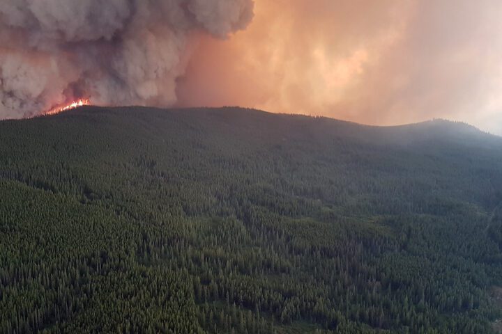 Aerial view of the Snowy Mountain wildfire burning along a forested ridge at night, with flames visible and smoke rising against a darkening sky over the dense forest landscape near Keremeos, British Columbia.