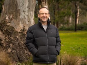 Adam Bandt smiling outdoors in front of a large gum tree, wearing a black jacket, after being announced as the next CEO of the Australian Conservation Foundation.