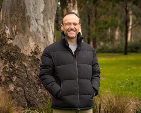 Adam Bandt smiling outdoors in front of a large gum tree, wearing a black jacket, after being announced as the next CEO of the Australian Conservation Foundation.