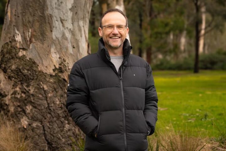 Adam Bandt smiling outdoors in front of a large gum tree, wearing a black jacket, after being announced as the next CEO of the Australian Conservation Foundation.