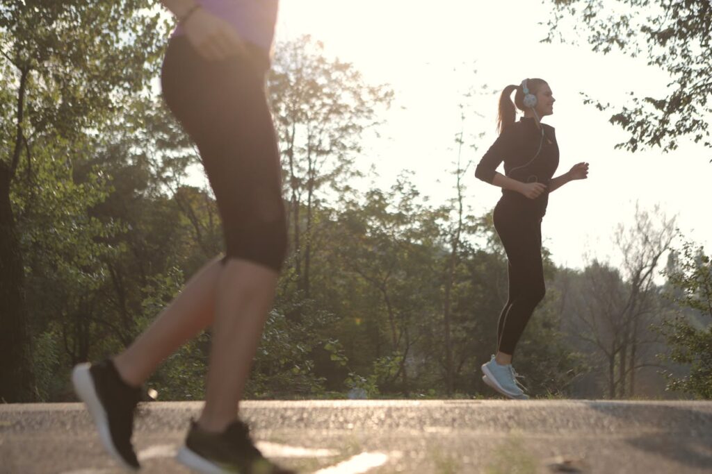 Two people jogging outdoors on a path surrounded by trees with sunlight filtering through the foliage.