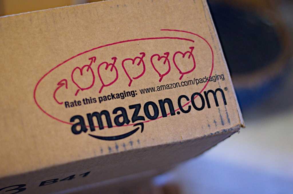 Amazon shipping box with five apple-shaped rating symbols circled in red and text saying "Rate this packaging: www.amazon.com/packaging" alongside the Amazon.com logo