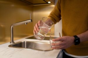 Representative Image. Person in yellow sweater pouring water from one clear glass to another at a kitchen sink with modern stainless steel faucet.
