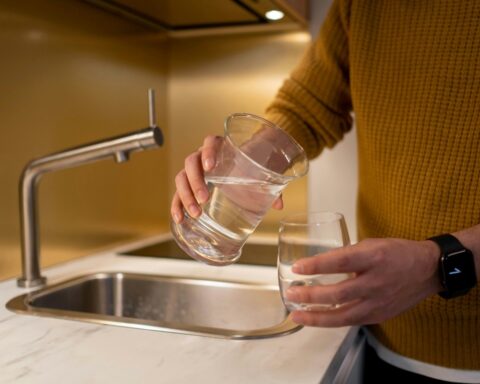 Representative Image. Person in yellow sweater pouring water from one clear glass to another at a kitchen sink with modern stainless steel faucet.