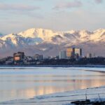 Downtown Anchorage, Alaska, with mid‑rise buildings reflecting in calm water, backed by snow‑covered mountains at sunrise or sunset.