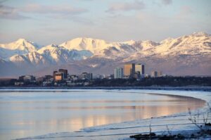 Downtown Anchorage, Alaska, with mid‑rise buildings reflecting in calm water, backed by snow‑covered mountains at sunrise or sunset.