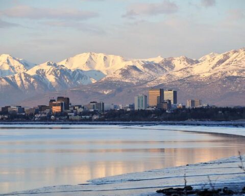 Downtown Anchorage, Alaska, with mid‑rise buildings reflecting in calm water, backed by snow‑covered mountains at sunrise or sunset.