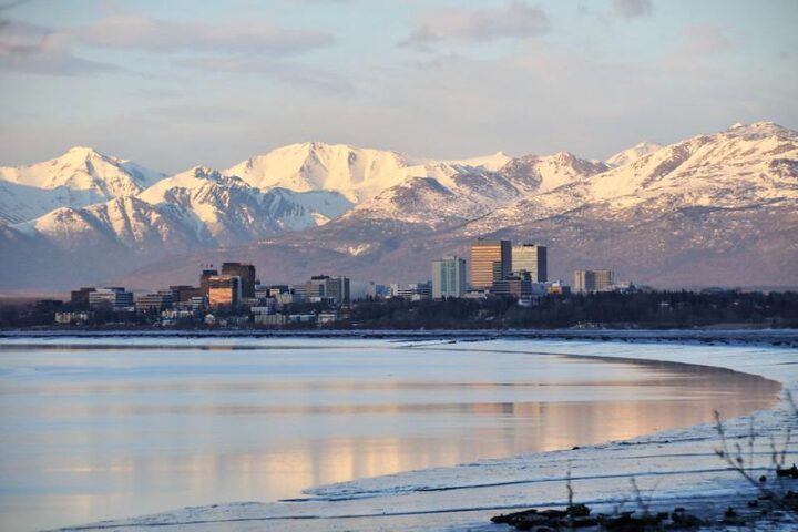 Downtown Anchorage, Alaska, with mid‑rise buildings reflecting in calm water, backed by snow‑covered mountains at sunrise or sunset.