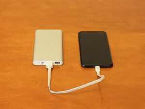 Two portable power banks - one white and one black - connected by a USB charging cable on an orange surface, similar to the recalled Anker models that pose fire hazards.