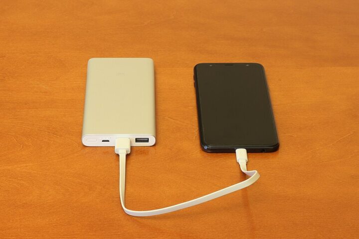 Two portable power banks - one white and one black - connected by a USB charging cable on an orange surface, similar to the recalled Anker models that pose fire hazards.