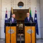 Anthony Albanese and Chris Bowen standing at podiums with microphones outside Parliament House, flanked by Australian flags, during a press event on climate policy.