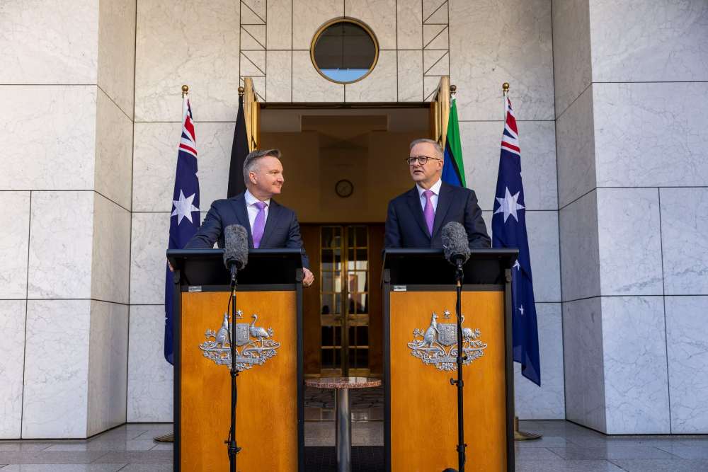 Anthony Albanese and Chris Bowen standing at podiums with microphones outside Parliament House, flanked by Australian flags, during a press event on climate policy.