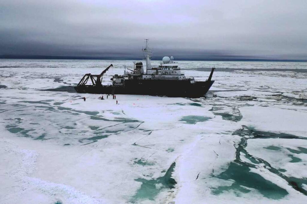 Research vessel Sikuliaq surrounded by Arctic ice floes with scientists conducting fieldwork on frozen surface during 2023 Chukchi Sea expedition to collect diatom samples from polar ice cores