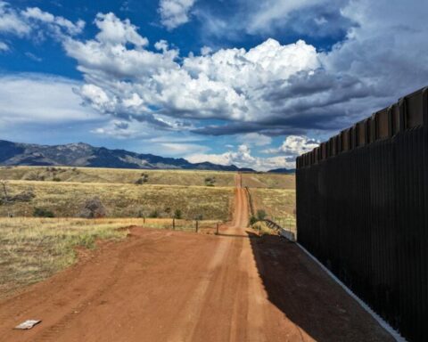 Border wall construction in San Rafael Valley showing a 30-foot black steel bollard barrier extending across grasslands with mountains and dramatic cloudy sky in the background.