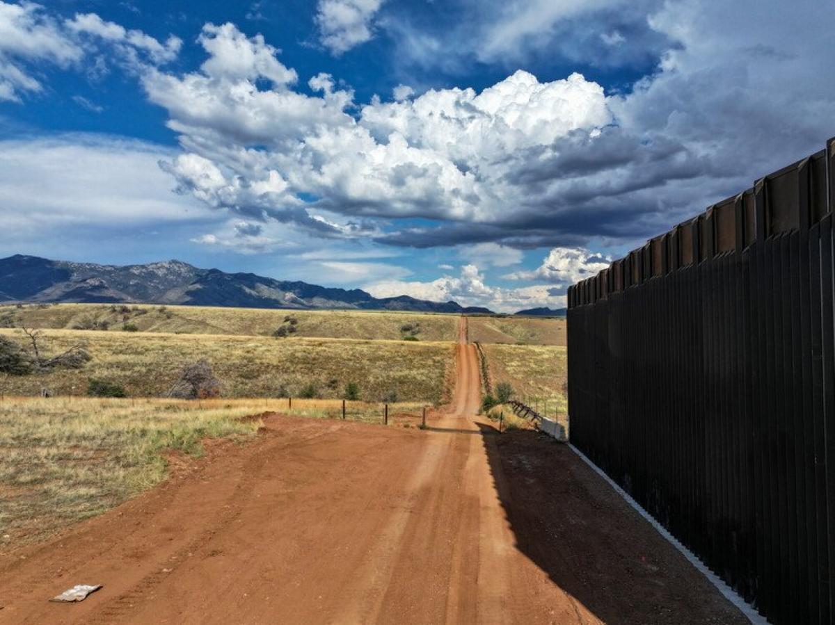 Border wall construction in San Rafael Valley showing a 30-foot black steel bollard barrier extending across grasslands with mountains and dramatic cloudy sky in the background.