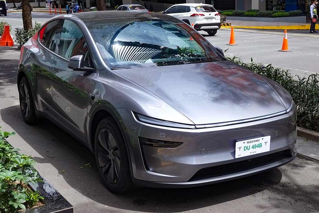 A 2025 Tesla Model Y in Quicksilver metallic color parked outdoors, showing the vehicle's streamlined front profile and aerodynamic design.