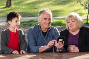 Older couple with grandson looking at mobile phone outdoors in park setting, smiling while checking information on smartphone screen.
