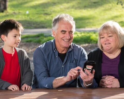 Older couple with grandson looking at mobile phone outdoors in park setting, smiling while checking information on smartphone screen.