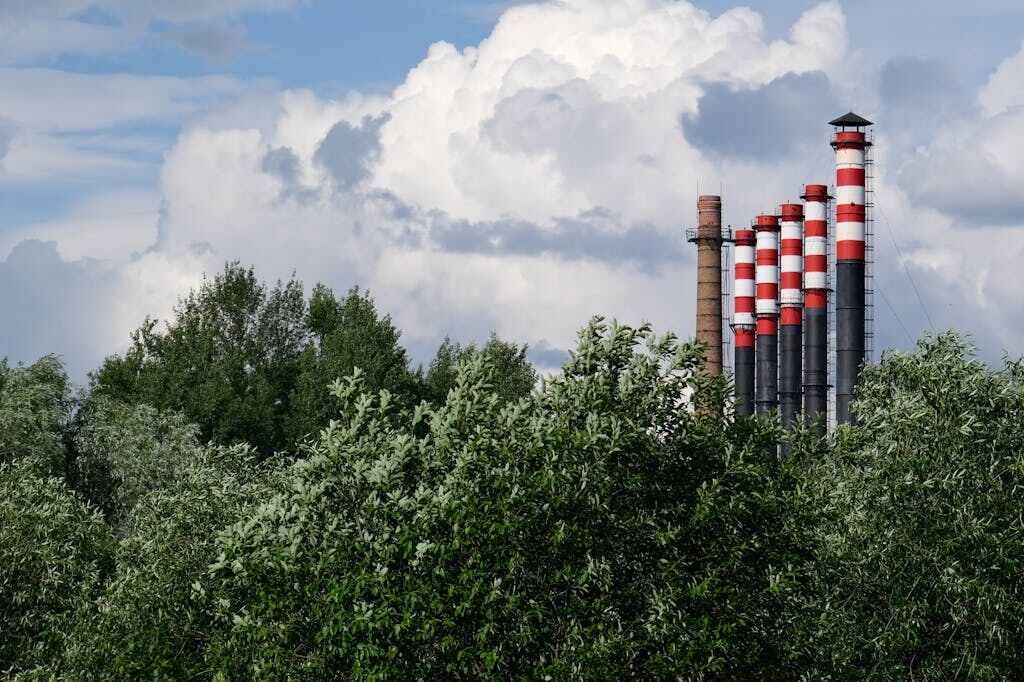 Industrial smokestacks with red and white striping rise above green trees against a cloudy blue sky, representing the challenge of reducing industrial emissions to meet climate targets.