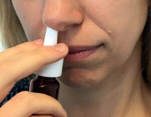 Person using a white nasal spray, holding the bottle with one hand and inserting the nozzle into their nostril with the other hand.