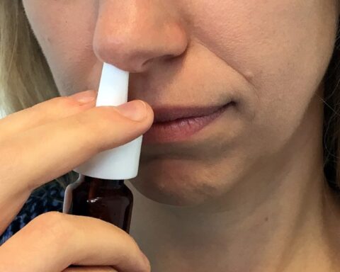 Person using a white nasal spray, holding the bottle with one hand and inserting the nozzle into their nostril with the other hand.