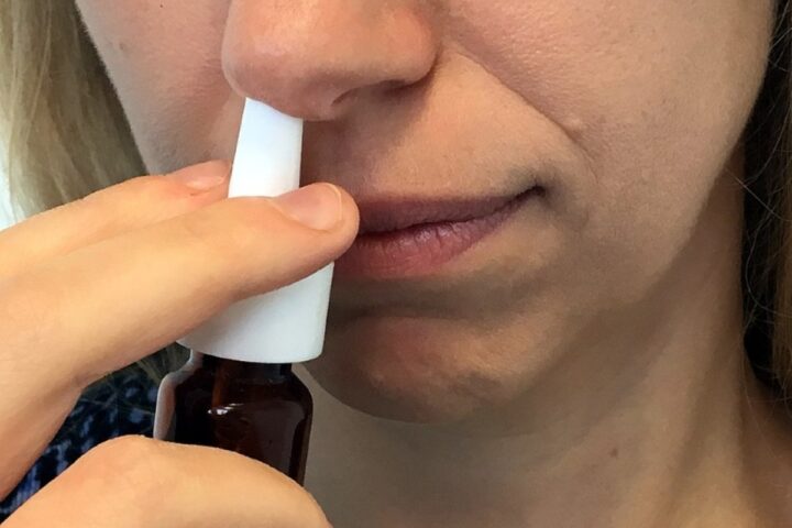 Person using a white nasal spray, holding the bottle with one hand and inserting the nozzle into their nostril with the other hand.