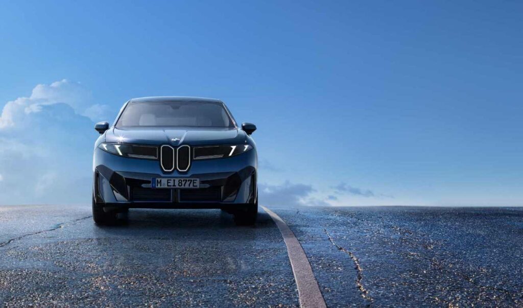 Front view of the blue 2026 BMW iX3 electric SUV on a wet road with blue sky in background, showcasing its distinctive new kidney grille design and sleek aerodynamic profile that's part of BMW's Neue Klasse design language.