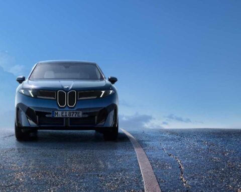 Front view of the blue 2026 BMW iX3 electric SUV on a wet road with blue sky in background, showcasing its distinctive new kidney grille design and sleek aerodynamic profile that's part of BMW's Neue Klasse design language.