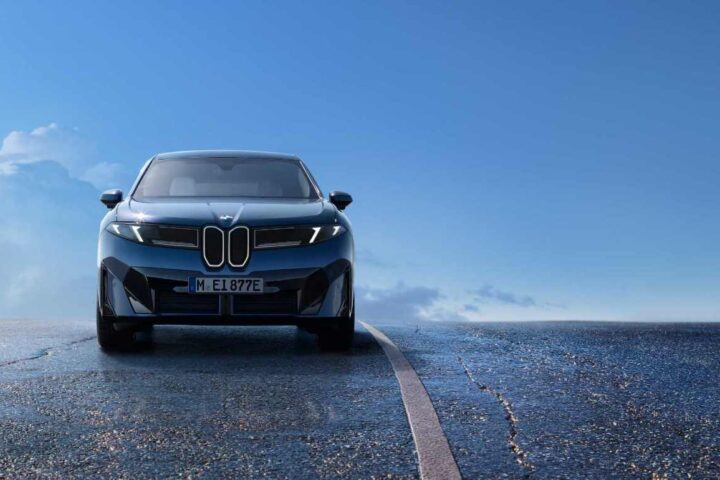 Front view of the blue 2026 BMW iX3 electric SUV on a wet road with blue sky in background, showcasing its distinctive new kidney grille design and sleek aerodynamic profile that's part of BMW's Neue Klasse design language.
