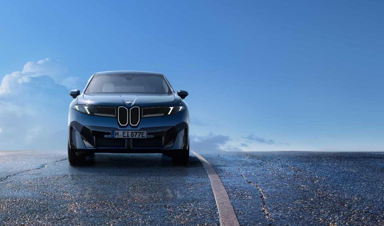 Front view of the blue 2026 BMW iX3 electric SUV on a wet road with blue sky in background, showcasing its distinctive new kidney grille design and sleek aerodynamic profile that's part of BMW's Neue Klasse design language.