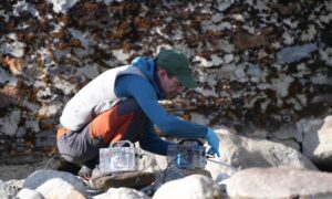 Researcher in blue jacket crouching by a rocky mountain stream, setting up transparent water filtration equipment to collect environmental DNA samples in Bhutan's Himalayan region.