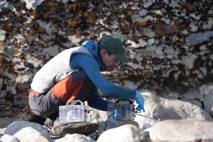 Researcher in blue jacket crouching by a rocky mountain stream, setting up transparent water filtration equipment to collect environmental DNA samples in Bhutan's Himalayan region.