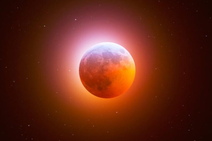 A long-exposure photograph of a total lunar eclipse, showing the moon glowing a deep, dramatic red against a starry night sky.