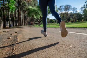 Person in blue jogging pants and white sneakers running on a path in a park with trees lining the sides, casting long shadows on the ground.