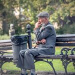 Elderly man in a gray suit and flat cap sitting alone on a park bench with a face mask pulled down, appearing thoughtful or concerned.